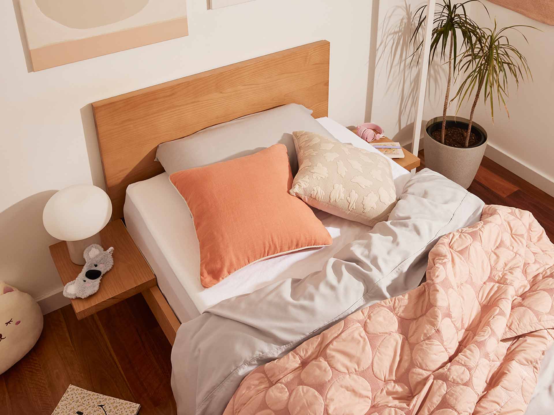 Bedroom with a wooden bed, light grey sheets, peach and beige cushions, and a peach quilt. A side table with a lamp and a potted plant nearby.