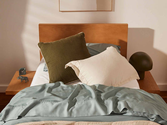 Bed with light wood headboard, adorned with olive and cream pillows. Sage green sheets, bedside table with lamp and decor.