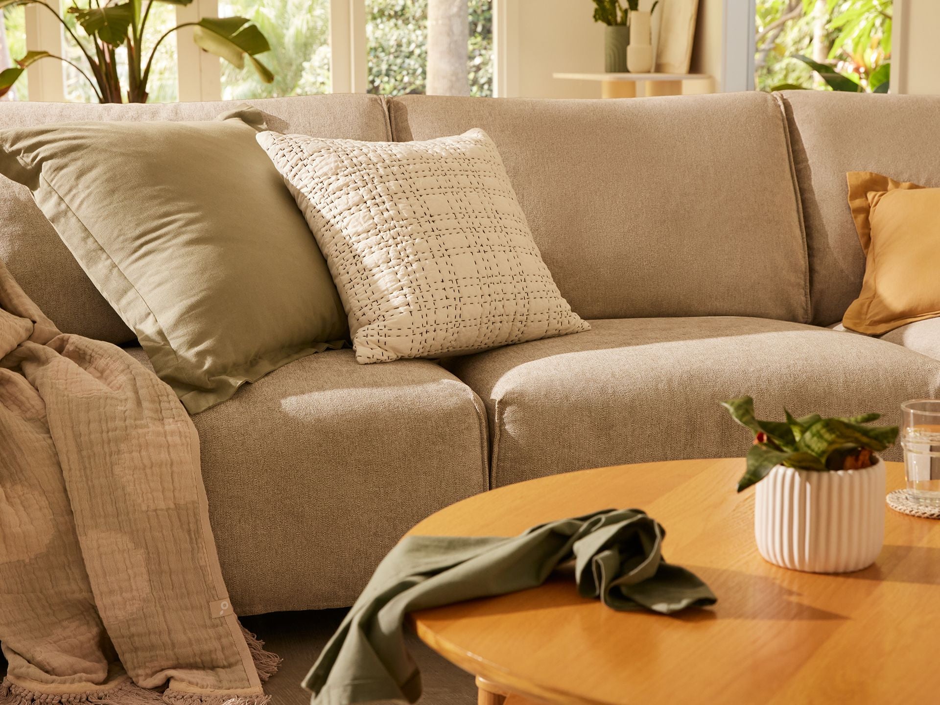A cosy living room with a light grey sectional sofa, green and patterned cushions, a wooden coffee table, and potted plants by the window.
