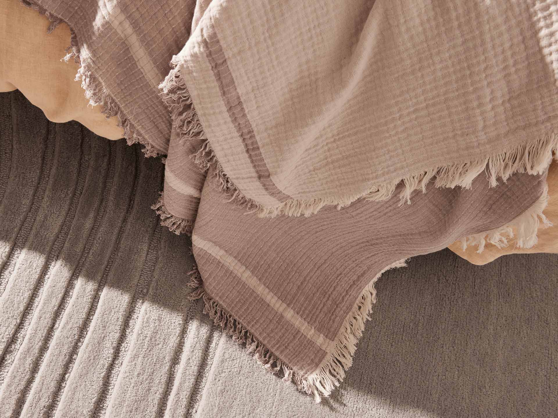 Beige textured throw blanket with fringed edges draped over a light brown surface. Subtle shadows create a cosy atmosphere.