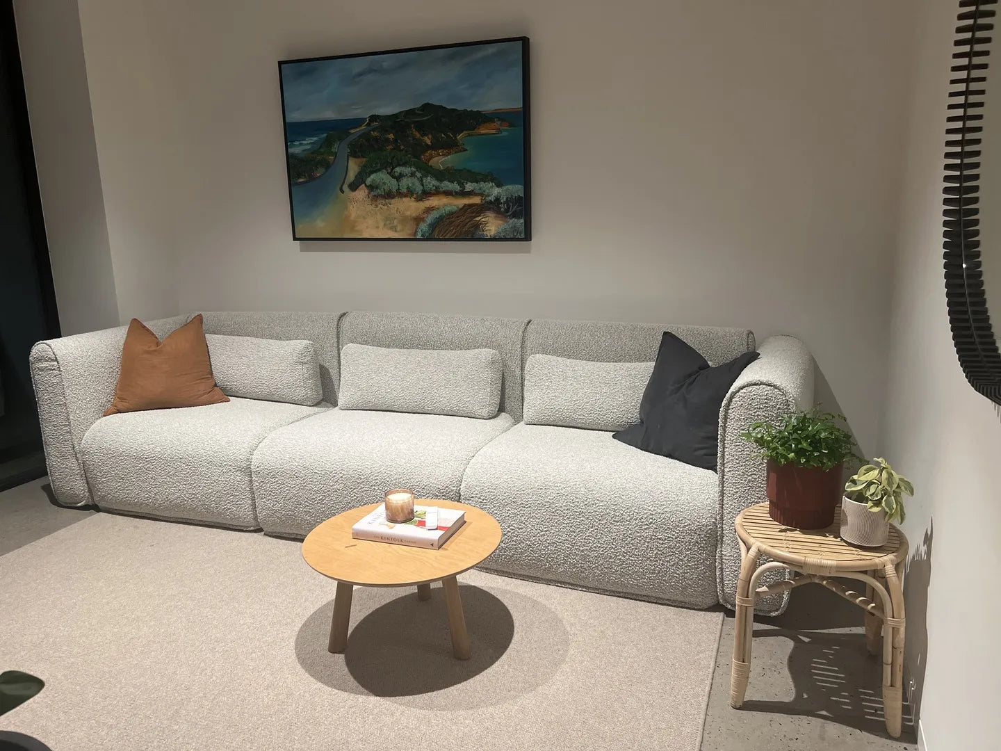 Light grey sectional sofa with orange and dark grey cushions in a cosy living room. A small round wooden table, art above, and plants nearby.
