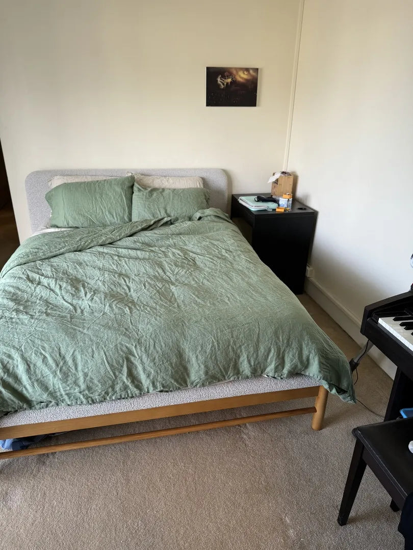 A bedroom with a bed featuring a green doona, grey upholstered headboard, and matching pillows. A black side table and keyboard are visible nearby.