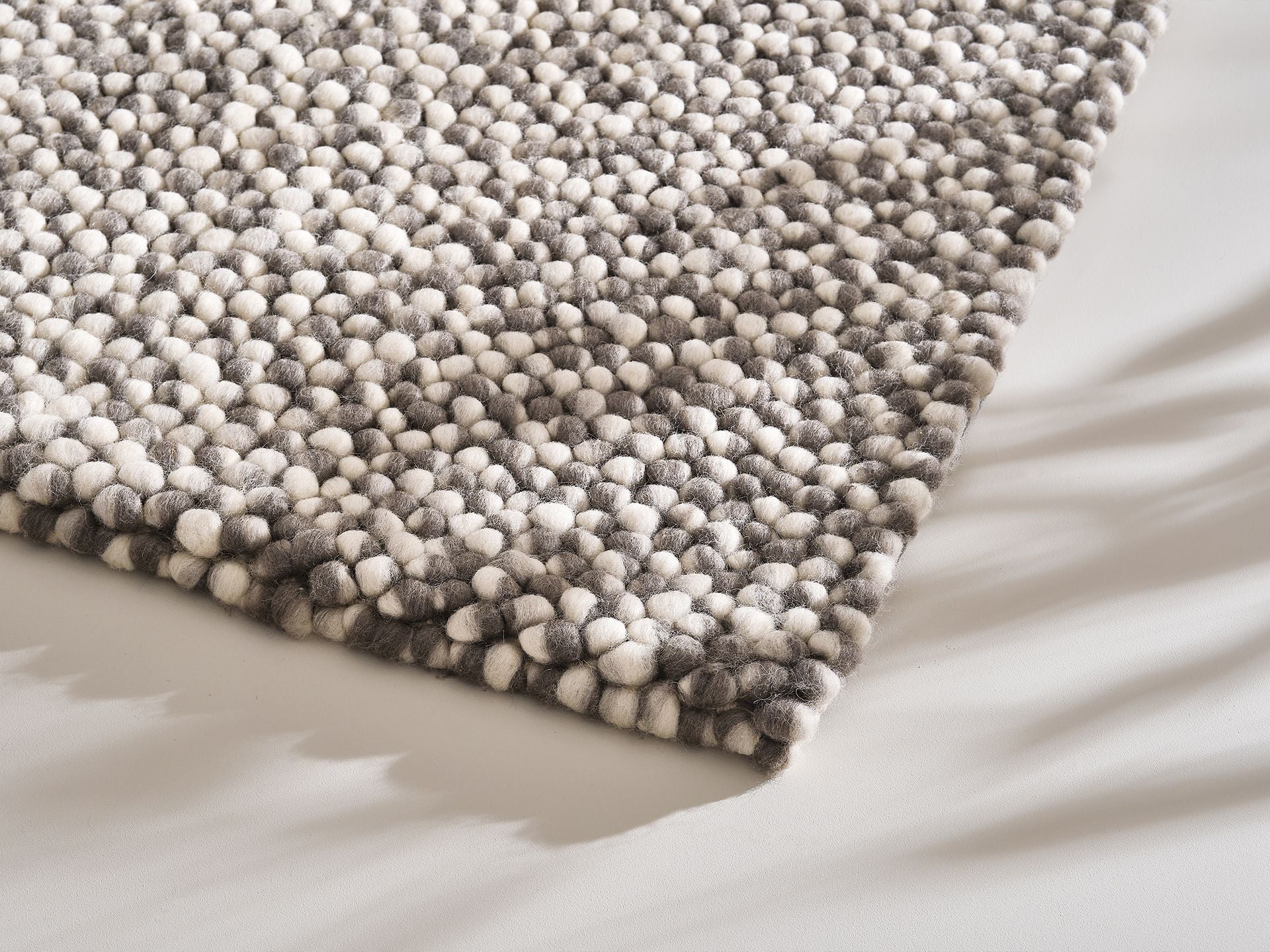 Close-up of a textured grey and white wool rug with a circular pattern on a light grey floor. Shadows cast along the surface.