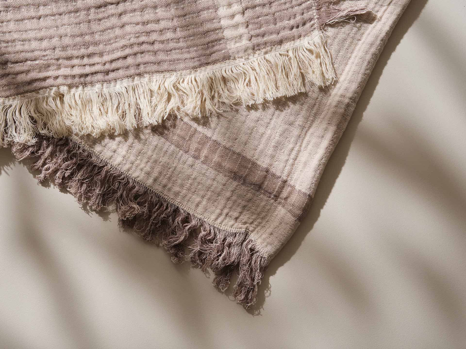 Close-up of a beige and white striped throw blanket with fringe, draped on a light surface. Long shadows suggest natural light.
