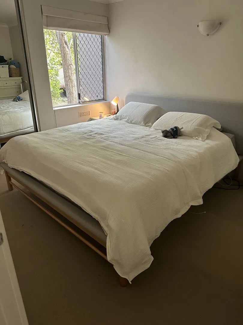 Bedroom with a large bed featuring a white quilt and pillows, grey upholstered headboard, bedside lamp, and window with a garden view.