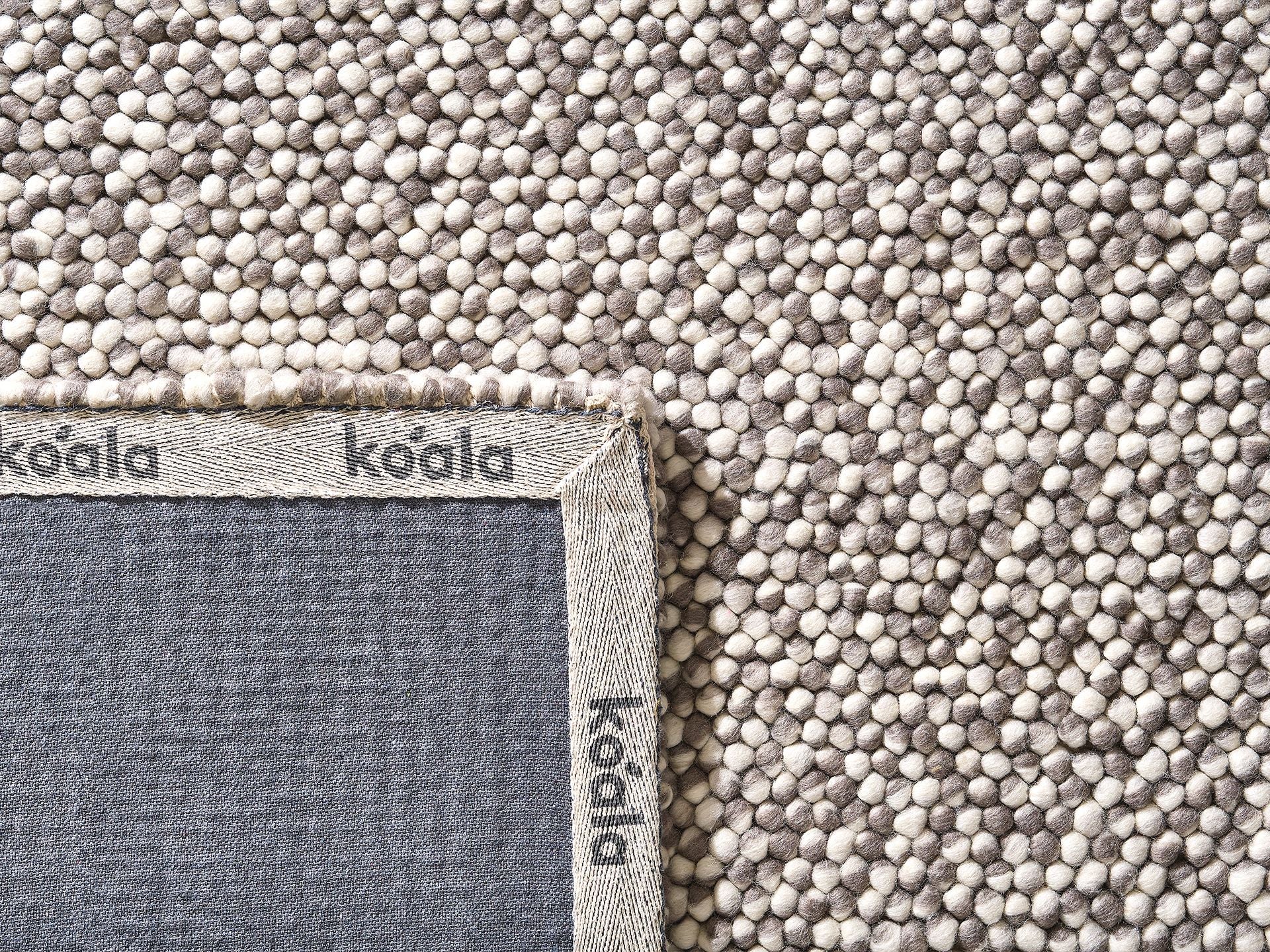 Close-up of a textured, grey and beige woven rug with a visible corner edge showing a grey fabric backing labeled "koala."