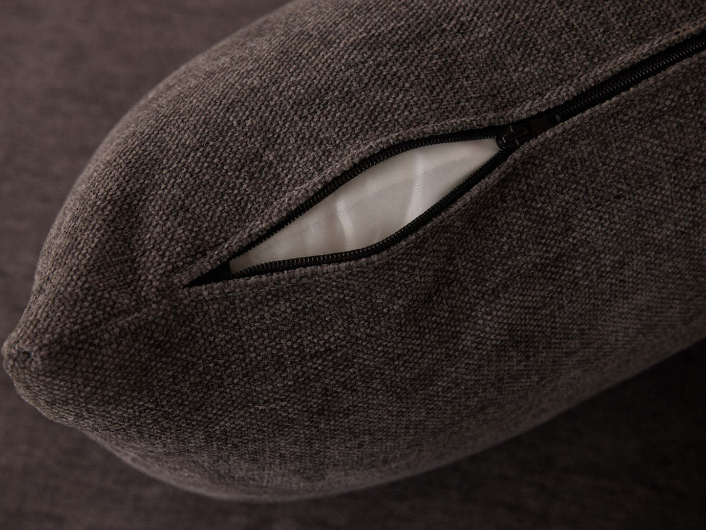 Dark grey cushion with a black zipper partially open, revealing white inner fabric. Close-up shot showcasing fabric texture.