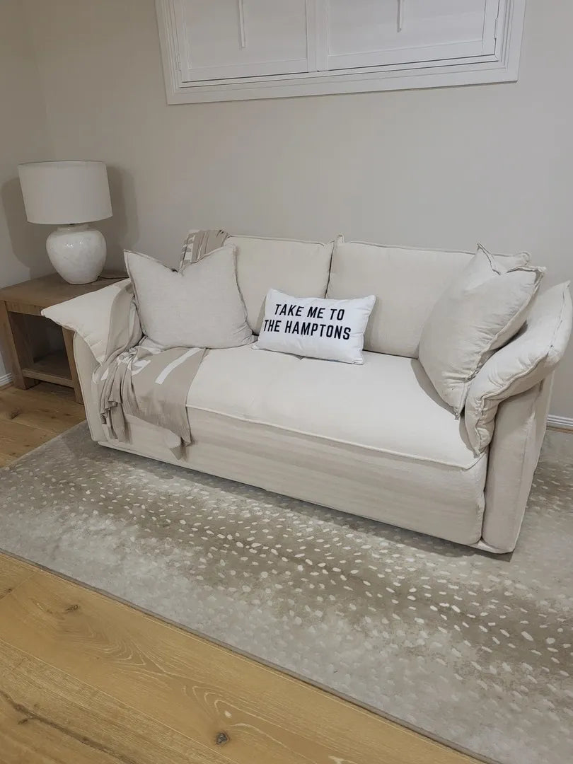 Cream sofa with cushy pillows, "Take Me to The Hamptons" cushion, and throw. Wooden side table with white lamp on beige patterned rug.