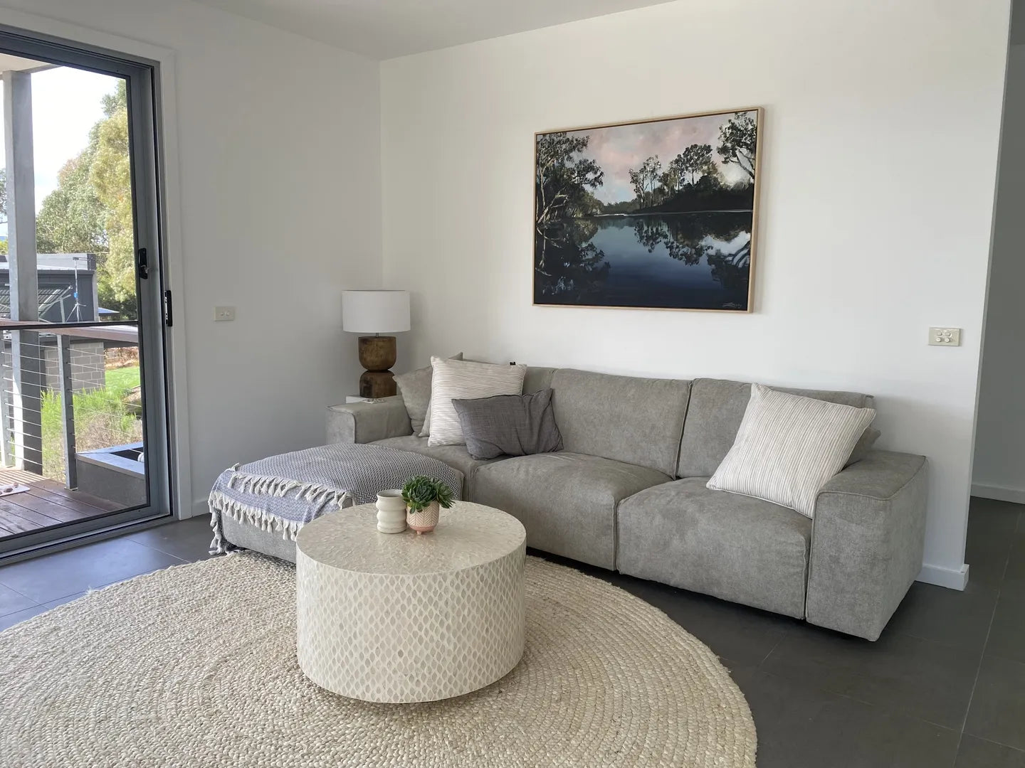 Living room with a light grey sectional sofa, round white coffee table, shaggy rug, and wall art. Sliding door to the left with garden view.