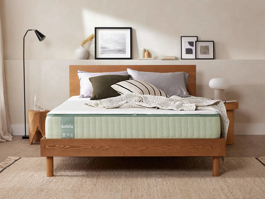 Wooden bed frame with a light green mattress, grey and white pillows, and a beige blanket. Artwork and lamps decorate the room.