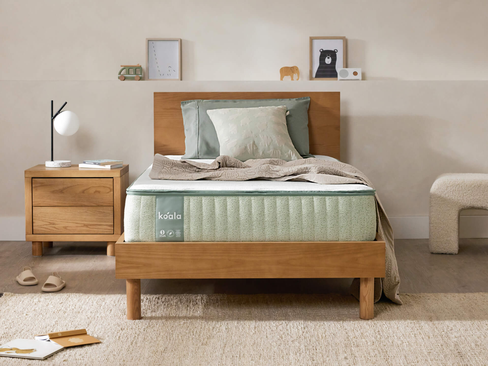 Bedroom scene with a wooden bed, green mattress, matching pillows. A wooden nightstand with a lamp and decor sits nearby. Neutral-toned decor.