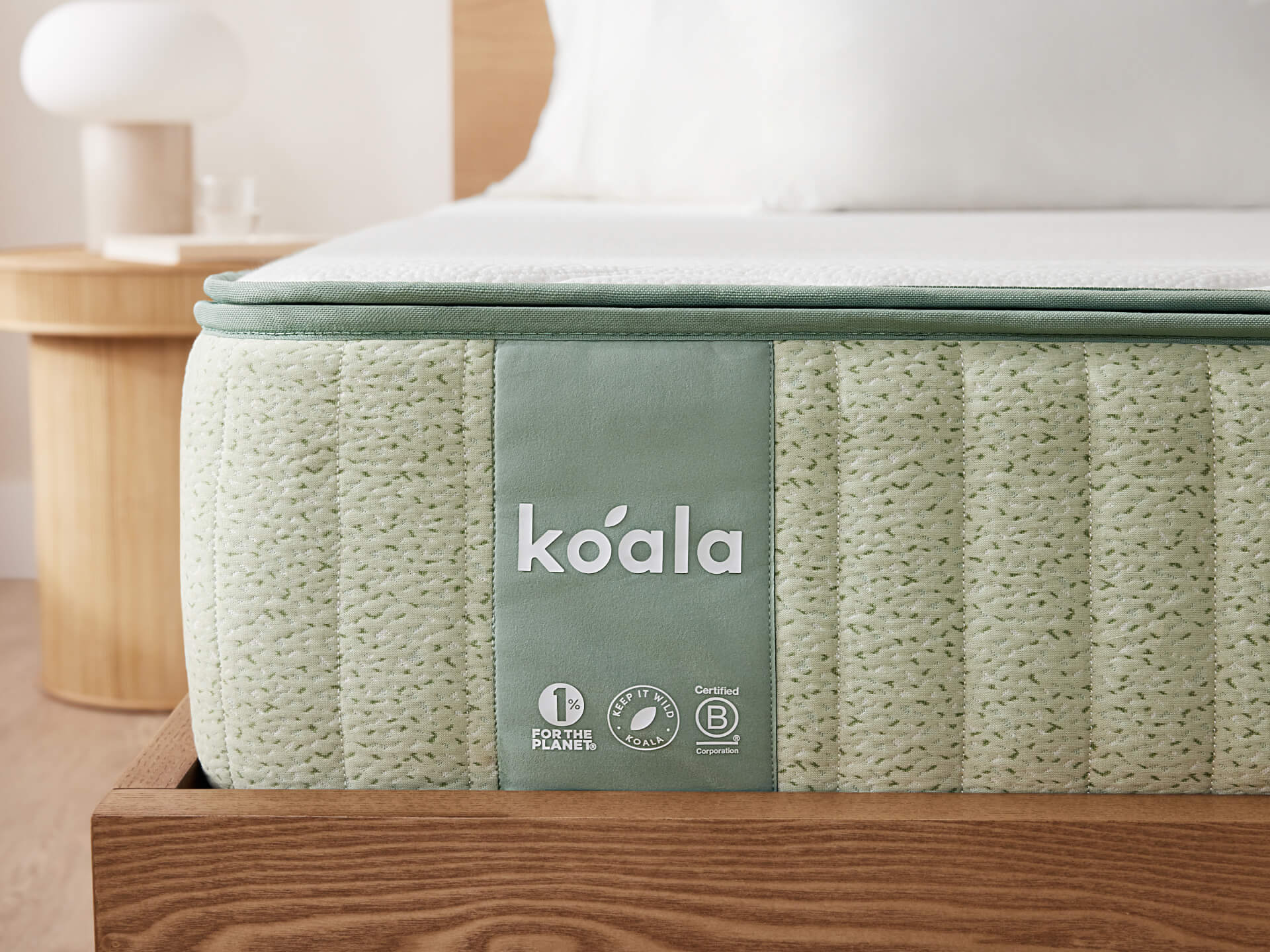 Light green mattress with "Koala" branding, on a wooden bed frame. Round wooden side table with a white lamp in the background.