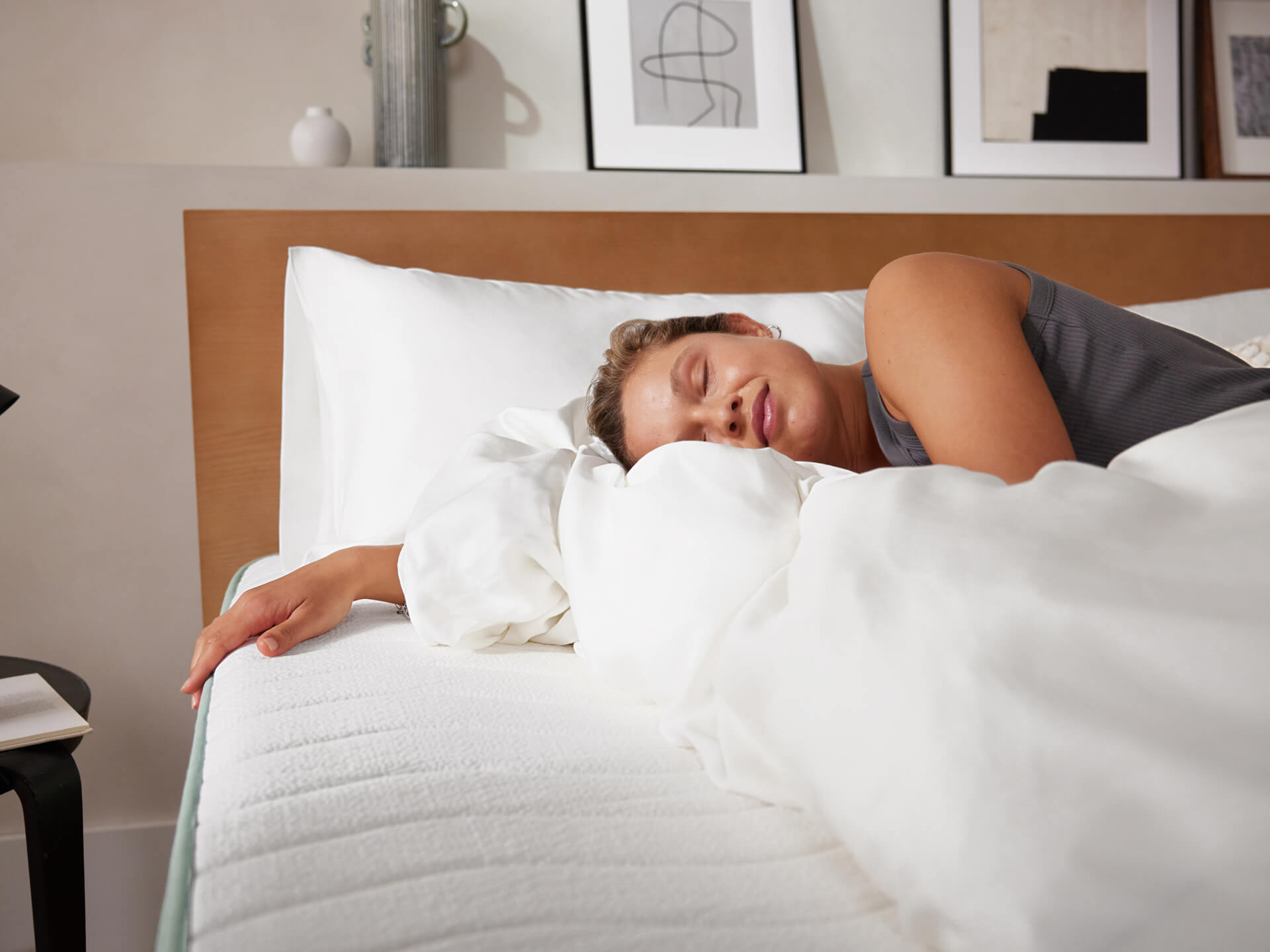 Woman sleeping on a white mattress with white bedding in a cosy bedroom. Wooden headboard and framed art in background.