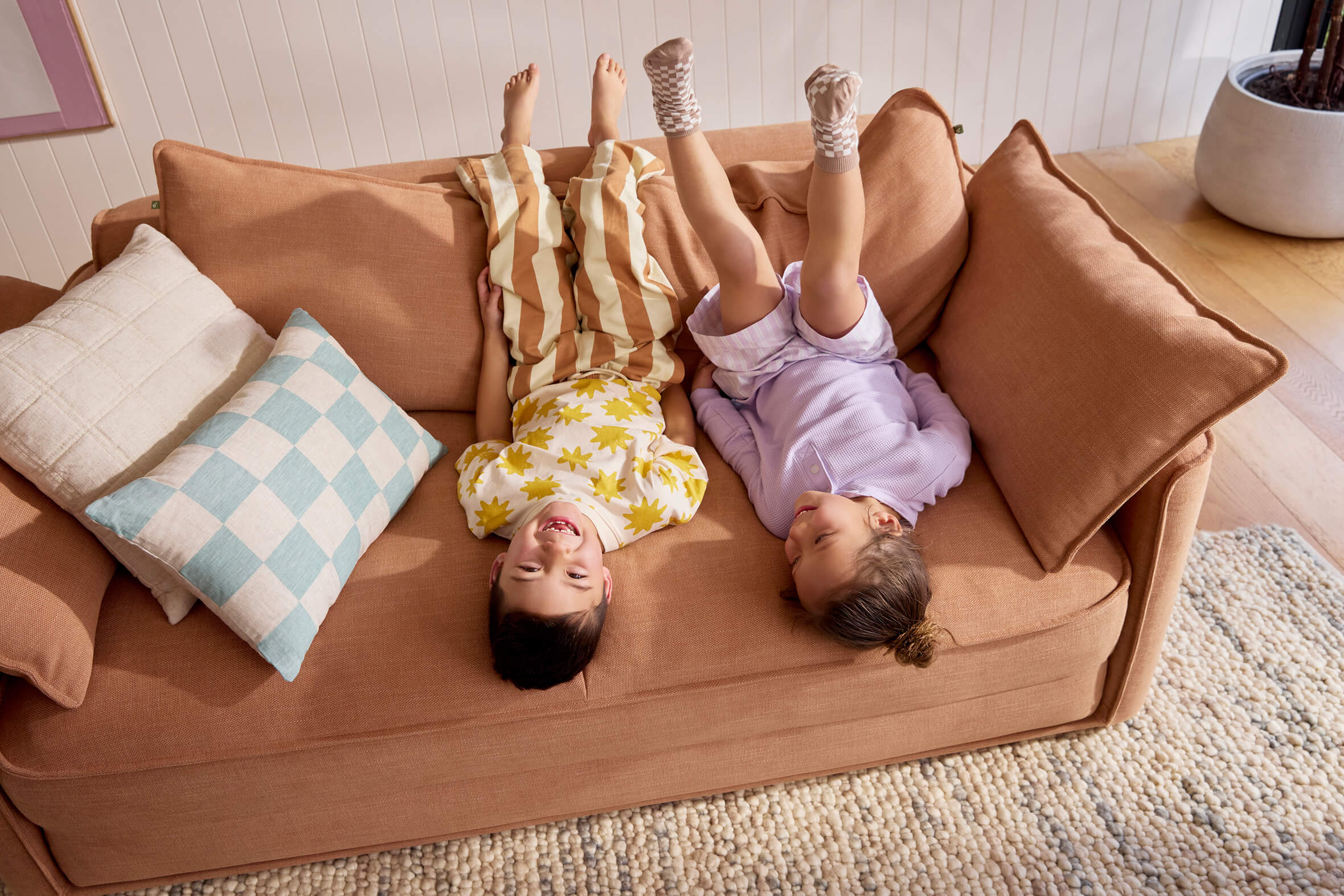 Two children playing upside down on terracotta Koala modular sofa with checkered blue and neutral scatter cushions