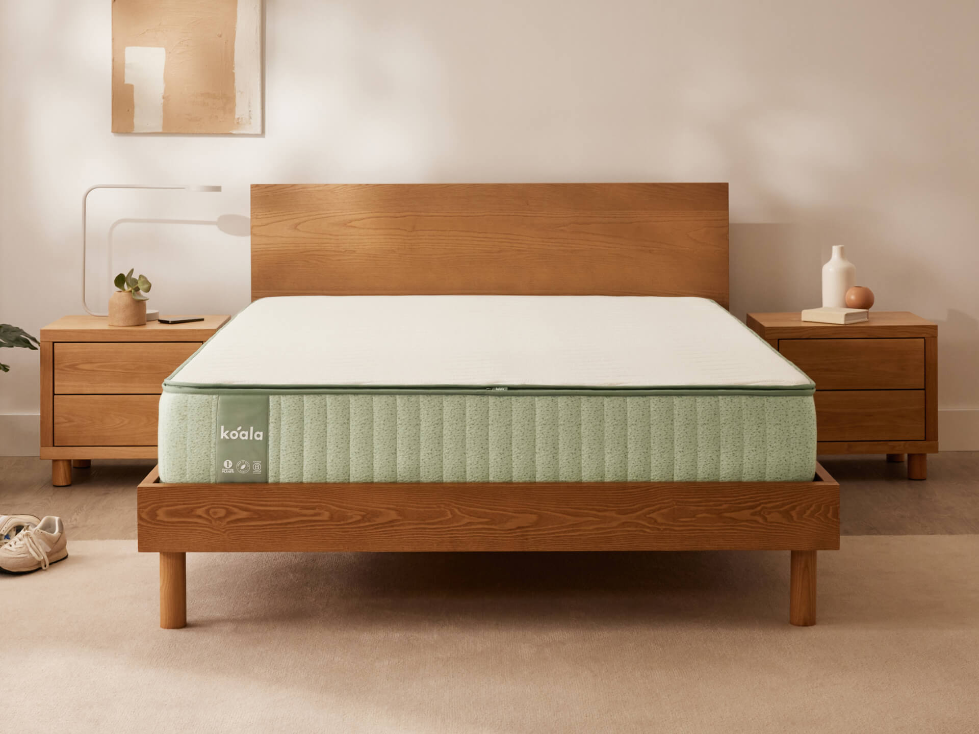 Wooden bed frame with a light green mattress in a bedroom. Two wooden bedside tables with decor items flank the bed. Neutral wall art above.