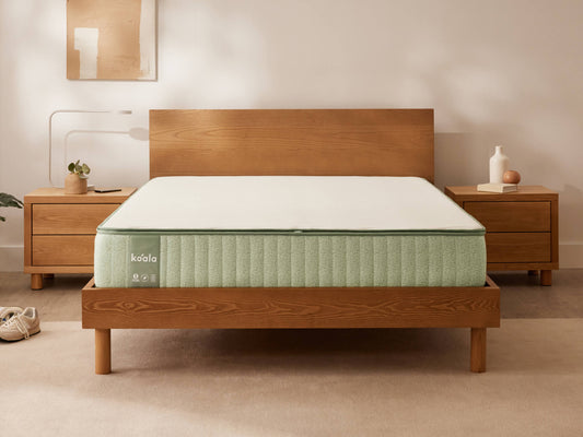 Wooden bed frame with a light green mattress in a bedroom. Two wooden bedside tables with decor items flank the bed. Neutral wall art above.