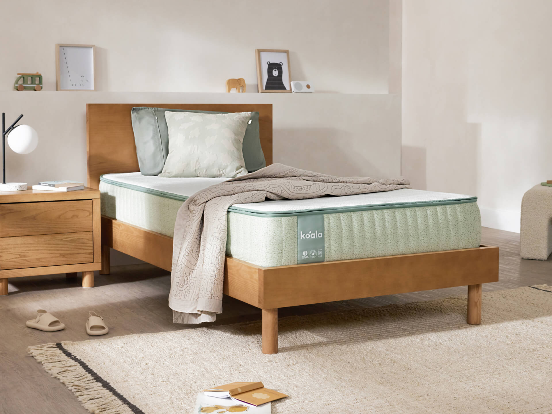 Bedroom with a light green mattress on a wooden bed frame, beige throw, wooden bedside table, and minimal decor on shelf.