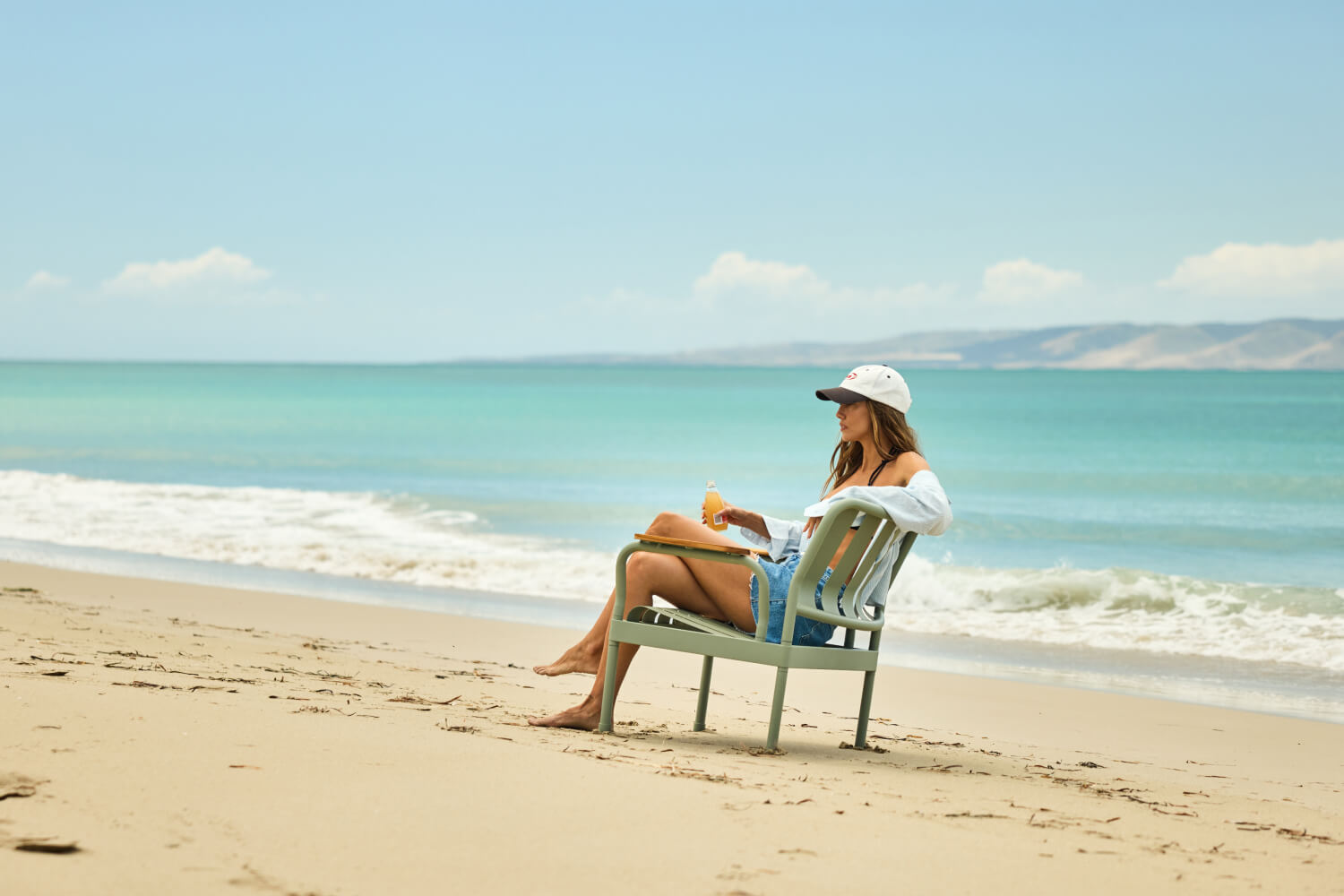 Person relaxing on a grey chair by the beach, wearing a white cap and light blue denim shorts, with waves and distant hills in the background.