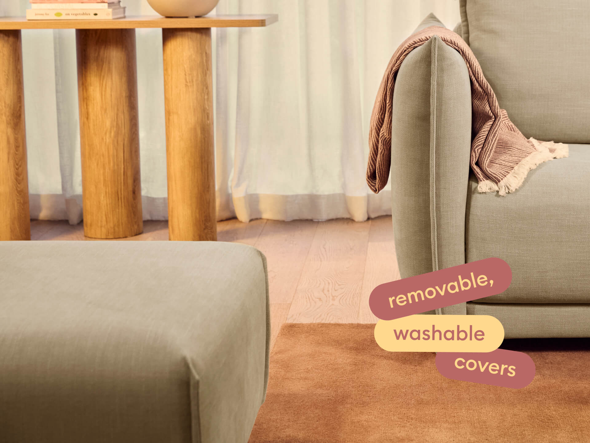 Beige sofa corner with a folded pink throw draped over the armrest next to a wooden table and ottoman on a brown rug