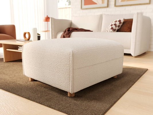 Cream-colored square ottoman with textured fabric and wooden legs placed on a brown rug in a bright living room.