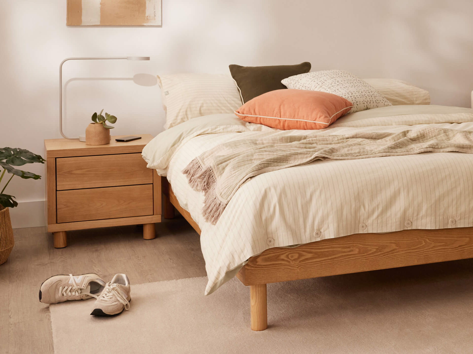 Cosy bedroom scene with a wooden bed frame, beige bedding, and assorted pillows. A wooden bedside table holds a lamp and plant. Shoes on floor.