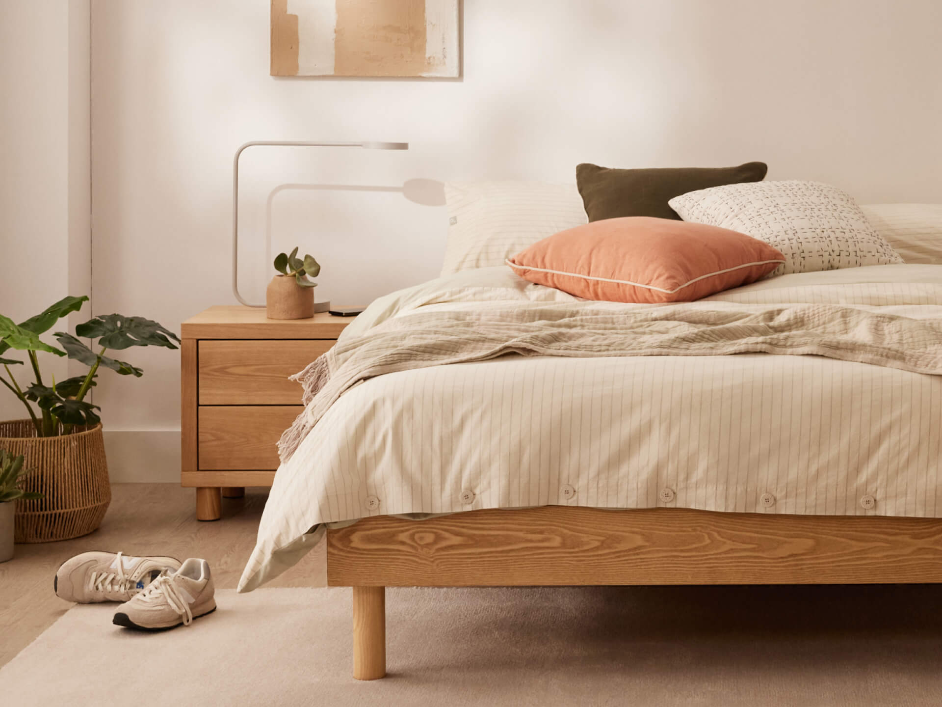 Bedroom with a light wood bed, white bedding, peach and dark cushions. A wooden bedside table holds a lamp and plant. Shoes and greenery nearby.
