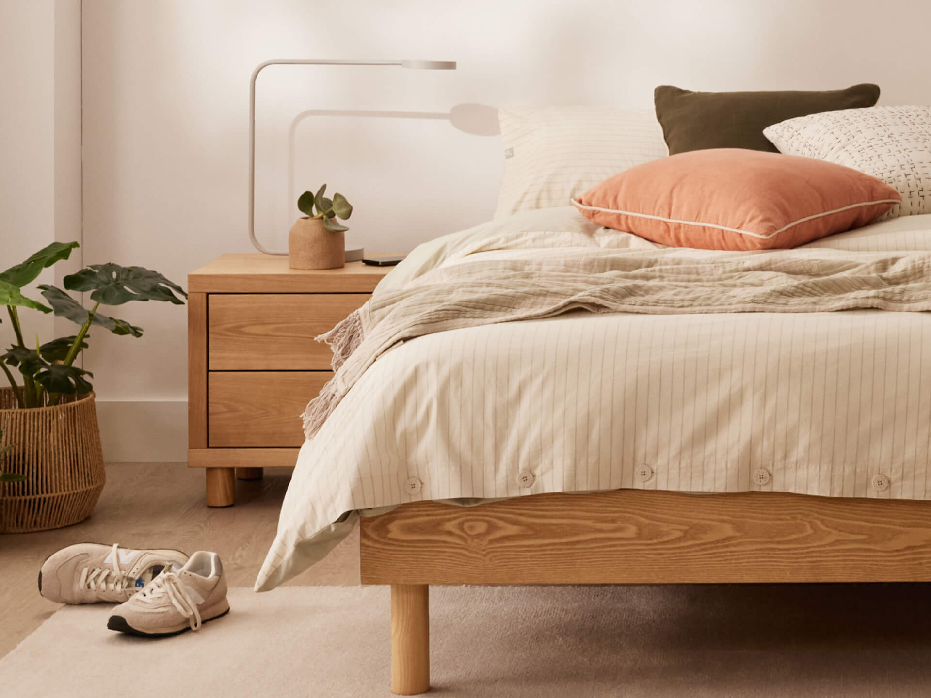 Modern bedroom with a light wood bed, bedside table, white lamp, potted plant, and neutral bedding. Sneakers on the floor add a casual touch.