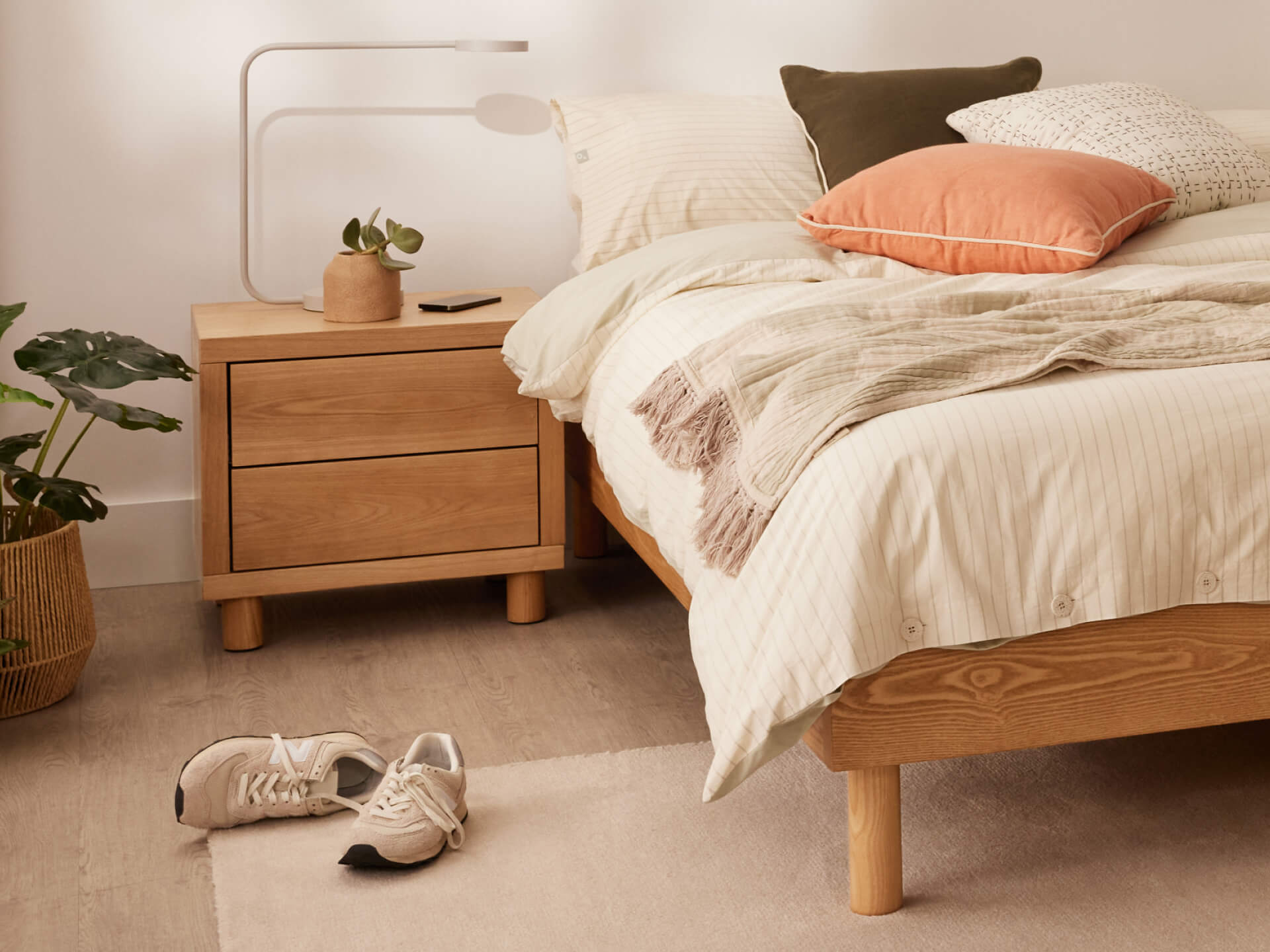 Bedroom with a light wooden bed and bedside table. Beige bedding, mixed pillows, indoor plant, sneakers on carpet, and modern lamp.