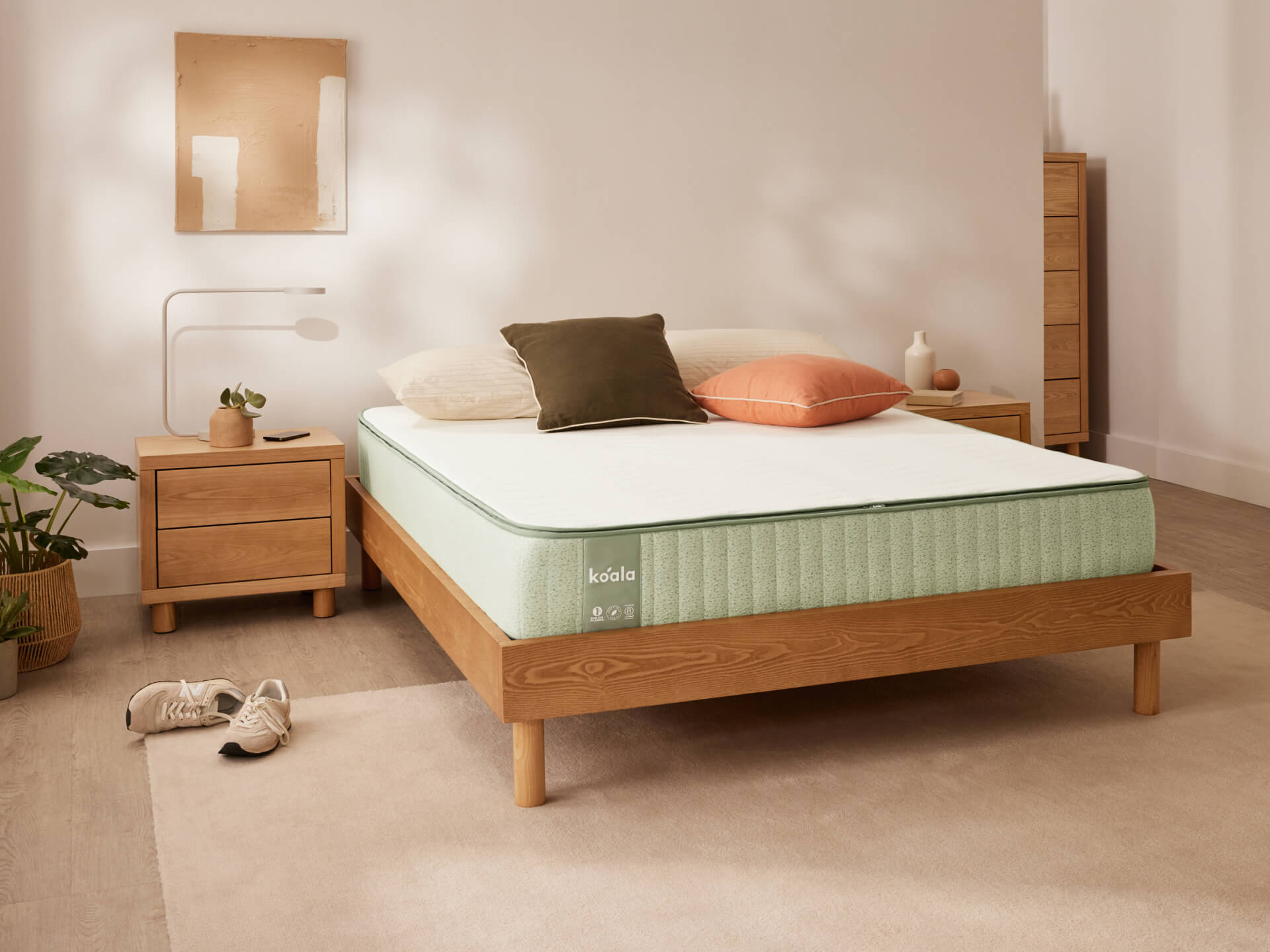 A bedroom with a wooden bed frame, light green mattress, assorted pillows, wooden side tables, indoor plant, and a neutral rug.