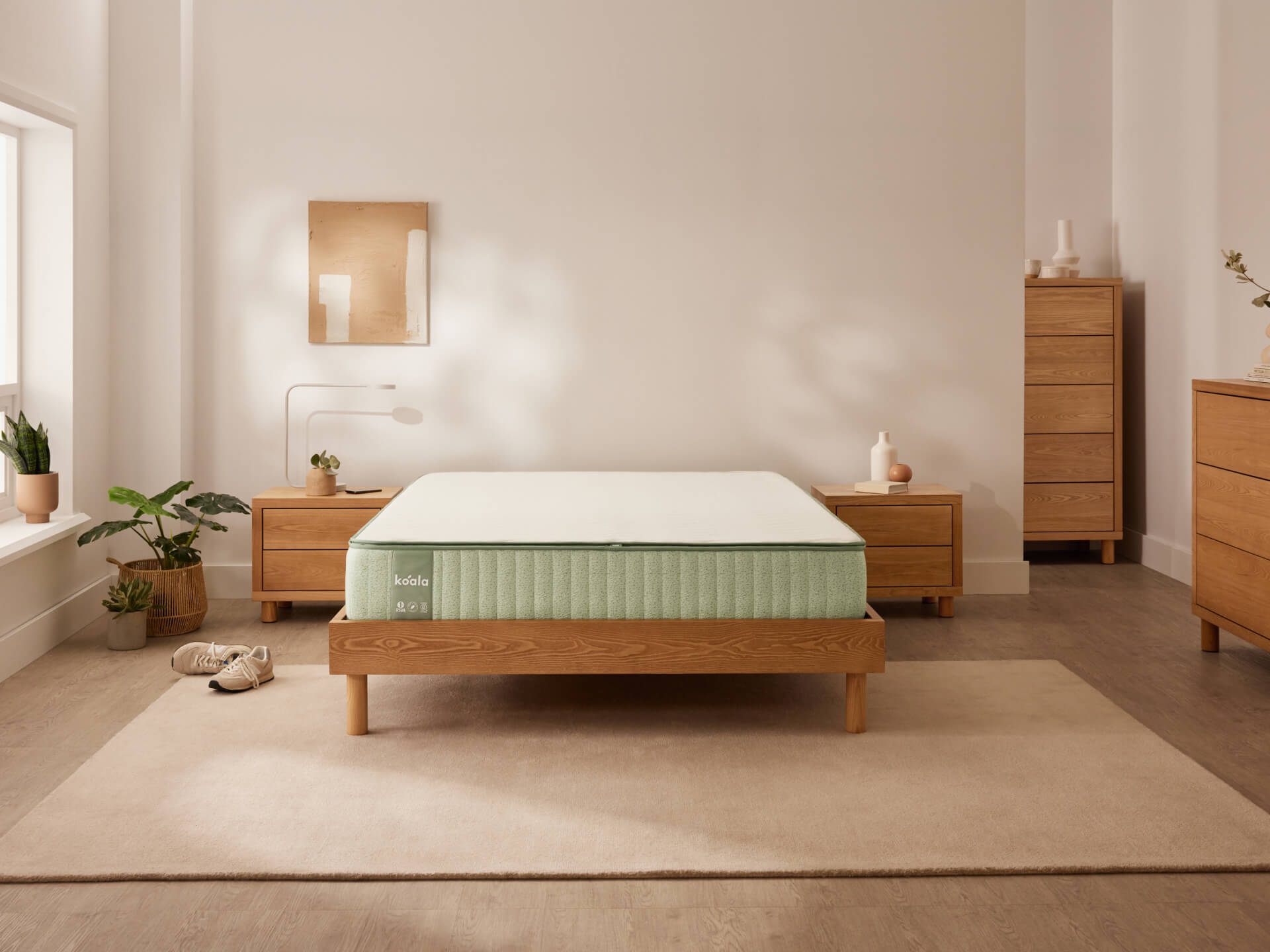 Minimalist bedroom with a wooden bed frame, mint-green mattress, light wood dressers, and plants. Beige rug and neutral walls.