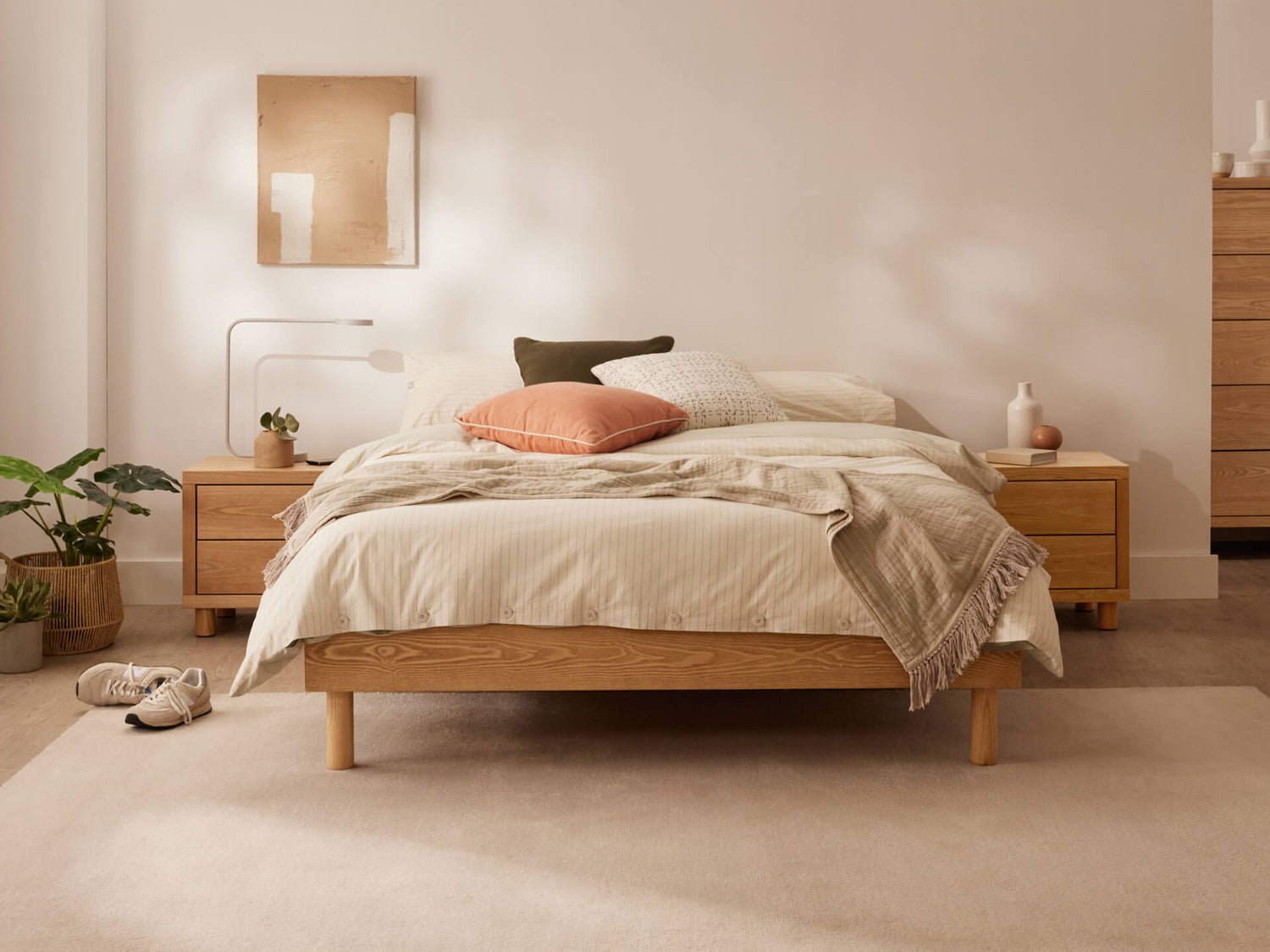 Beige bedroom with wooden bed, matching bedside tables, and soft lighting. Includes green plant, wall art, and a rug. Minimalist decor.