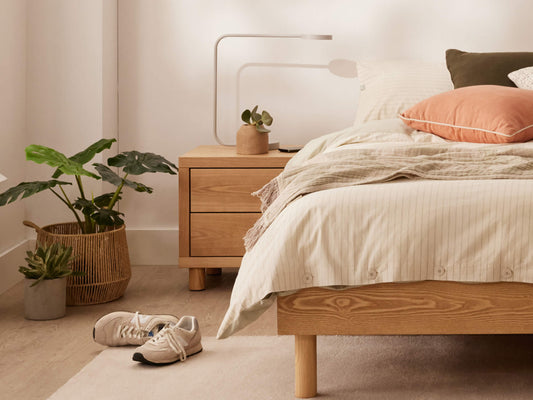 A cosy bedroom with a wooden bed, cream bedding, orange and olive cushions, a wooden nightstand with a plant, and sneakers on the floor.