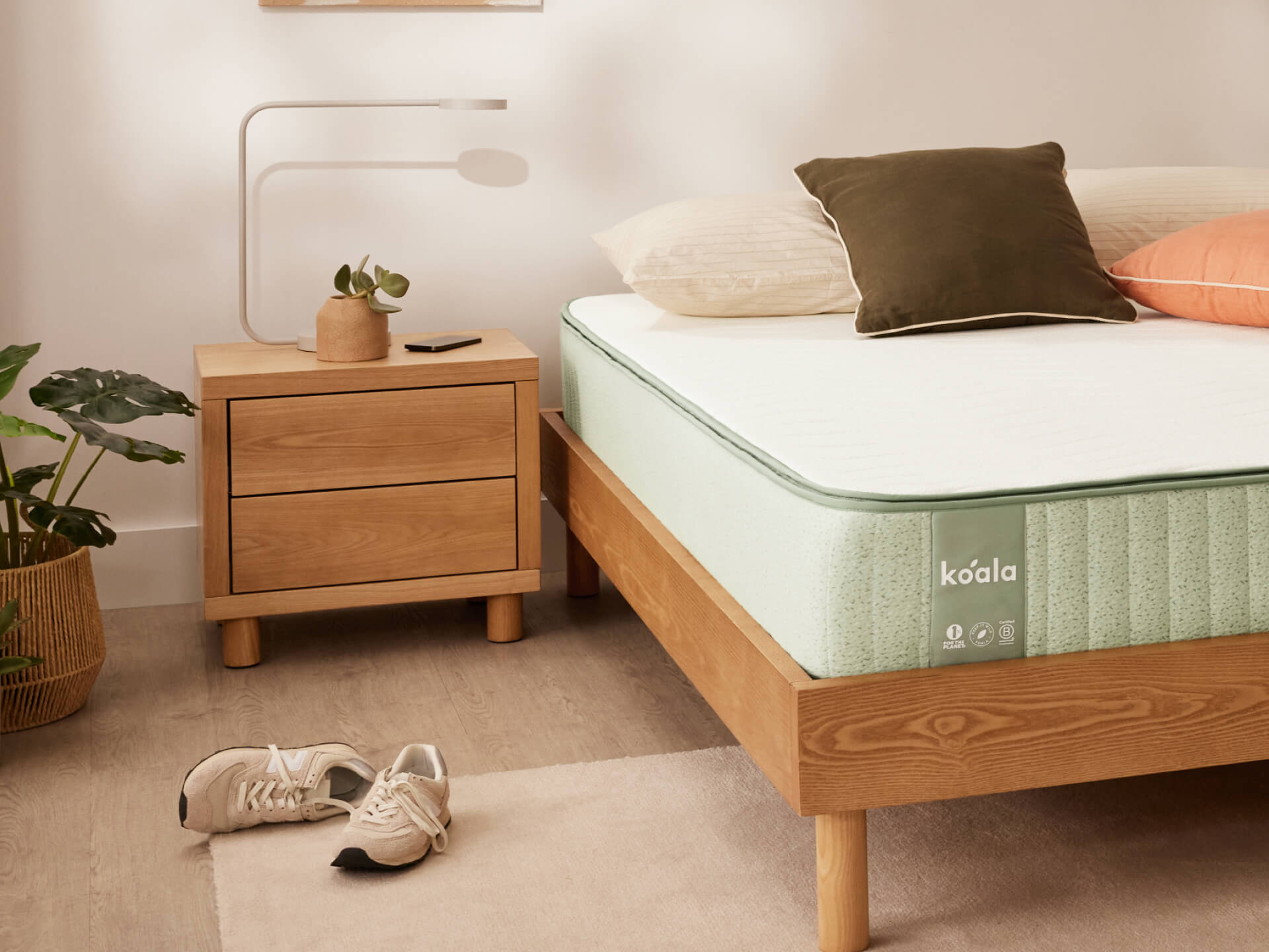 Bedroom with a light wood bed frame, mint green mattress, wooden bedside table, minimalist lamp, and plants. Shoes on light wood floor.