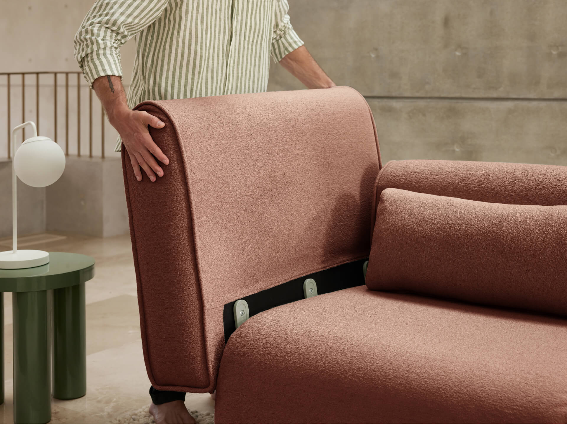 Person adjusting a modular sofa in muted pink fabric. Nearby, a round green side table with a white lamp on top. Modern interior setting.