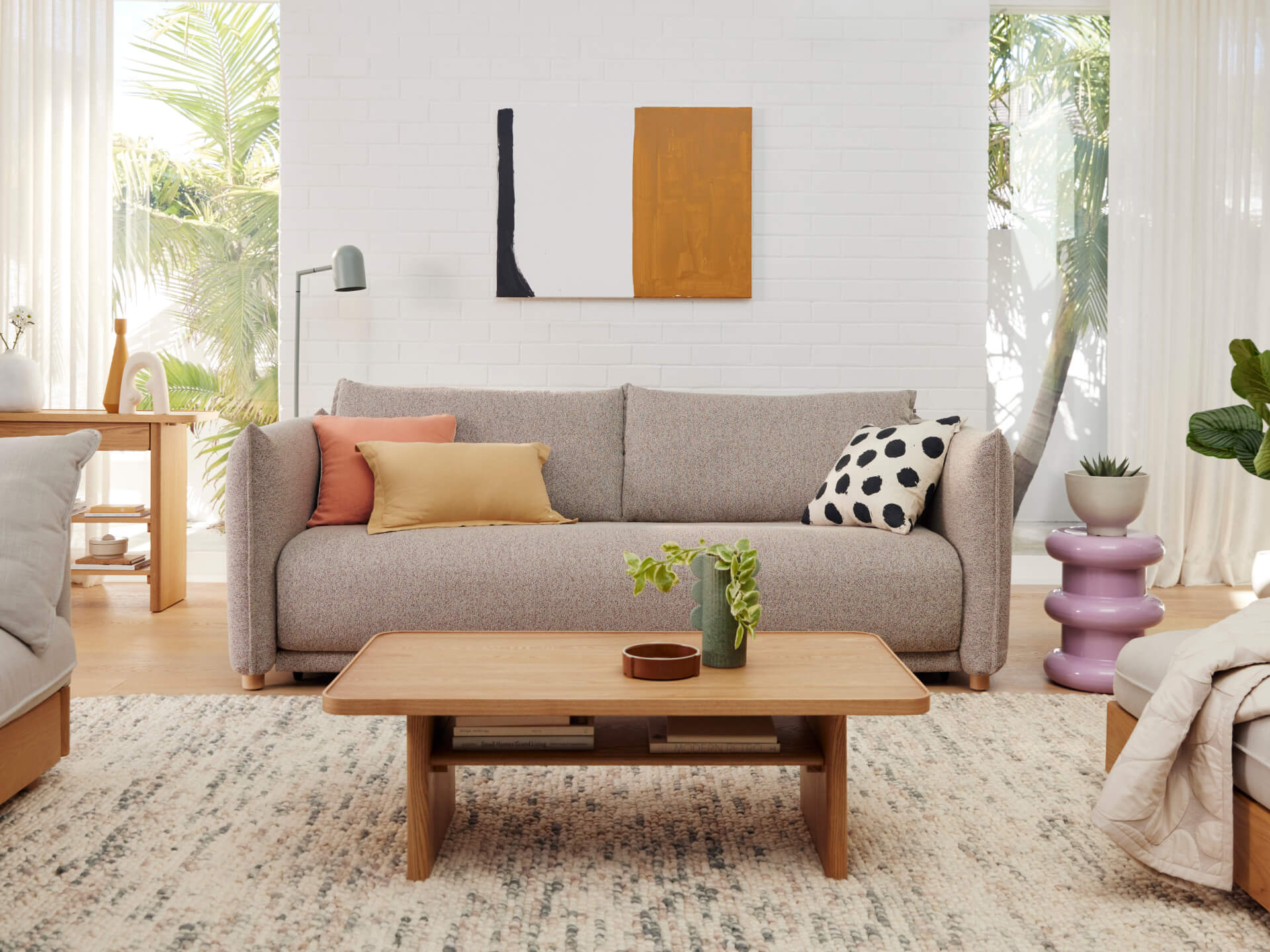 Beige couch with assorted cushions, wooden coffee table, plants, and abstract art in a modern living room. Light grey patterned rug.