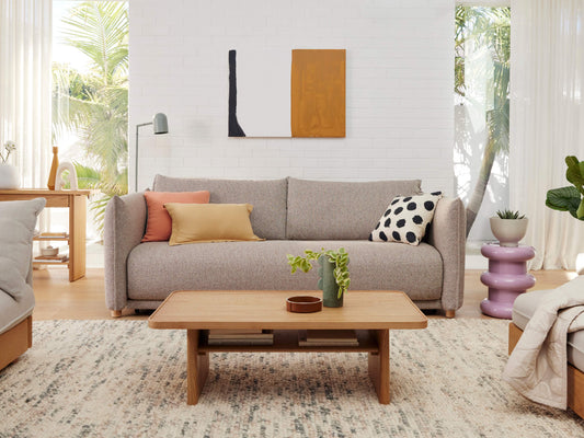 Beige couch with assorted cushions, wooden coffee table, plants, and abstract art in a modern living room. Light grey patterned rug.