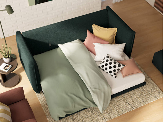 Green fabric daybed with a light green duvet, multi-coloured cushions, and a polka dot pillow on a cream rug in a living room setting.