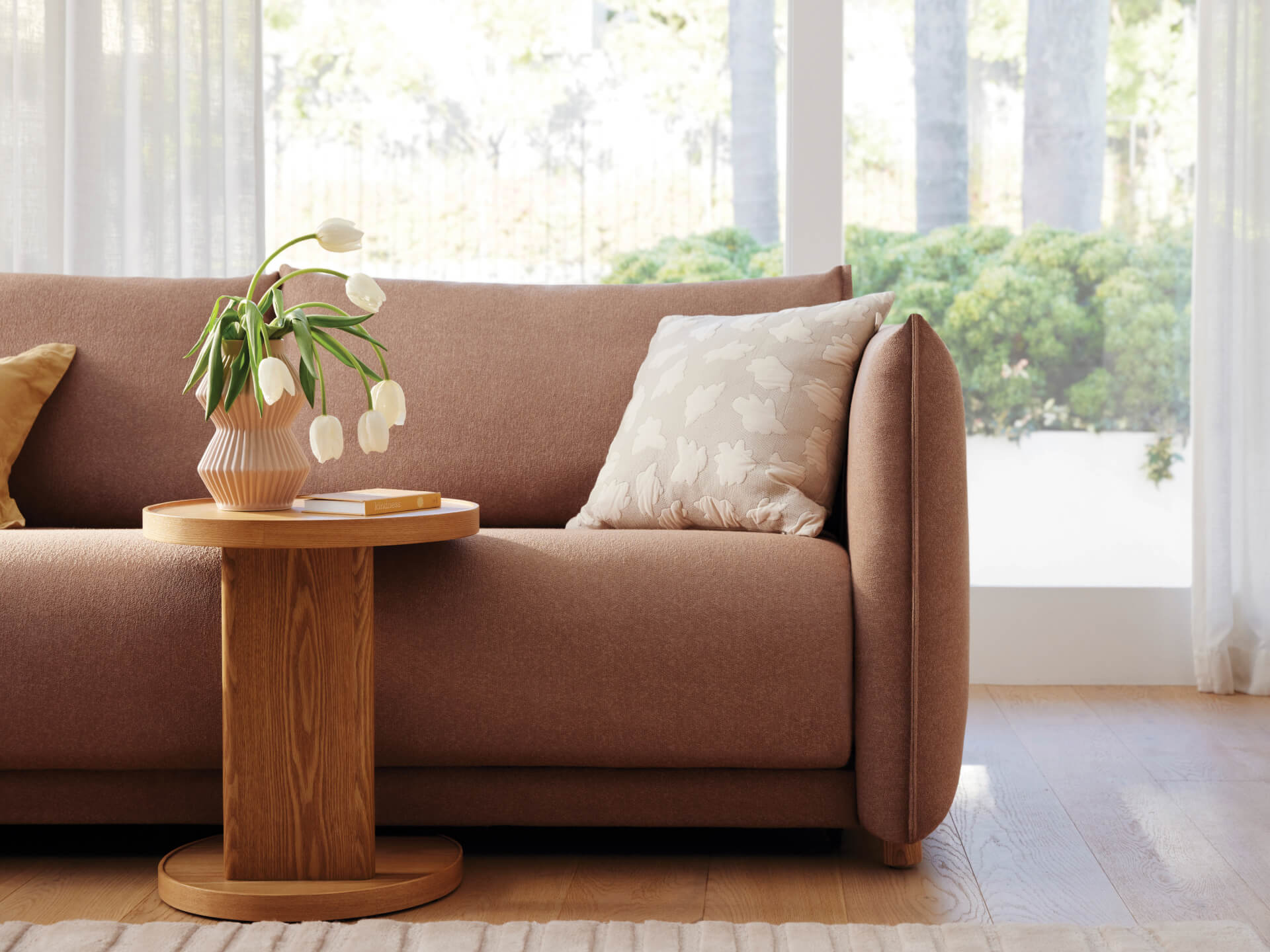 Modern living room with a soft pink sofa, beige and white cushions, a wooden side table, and a vase of white tulips. Large windows in background.