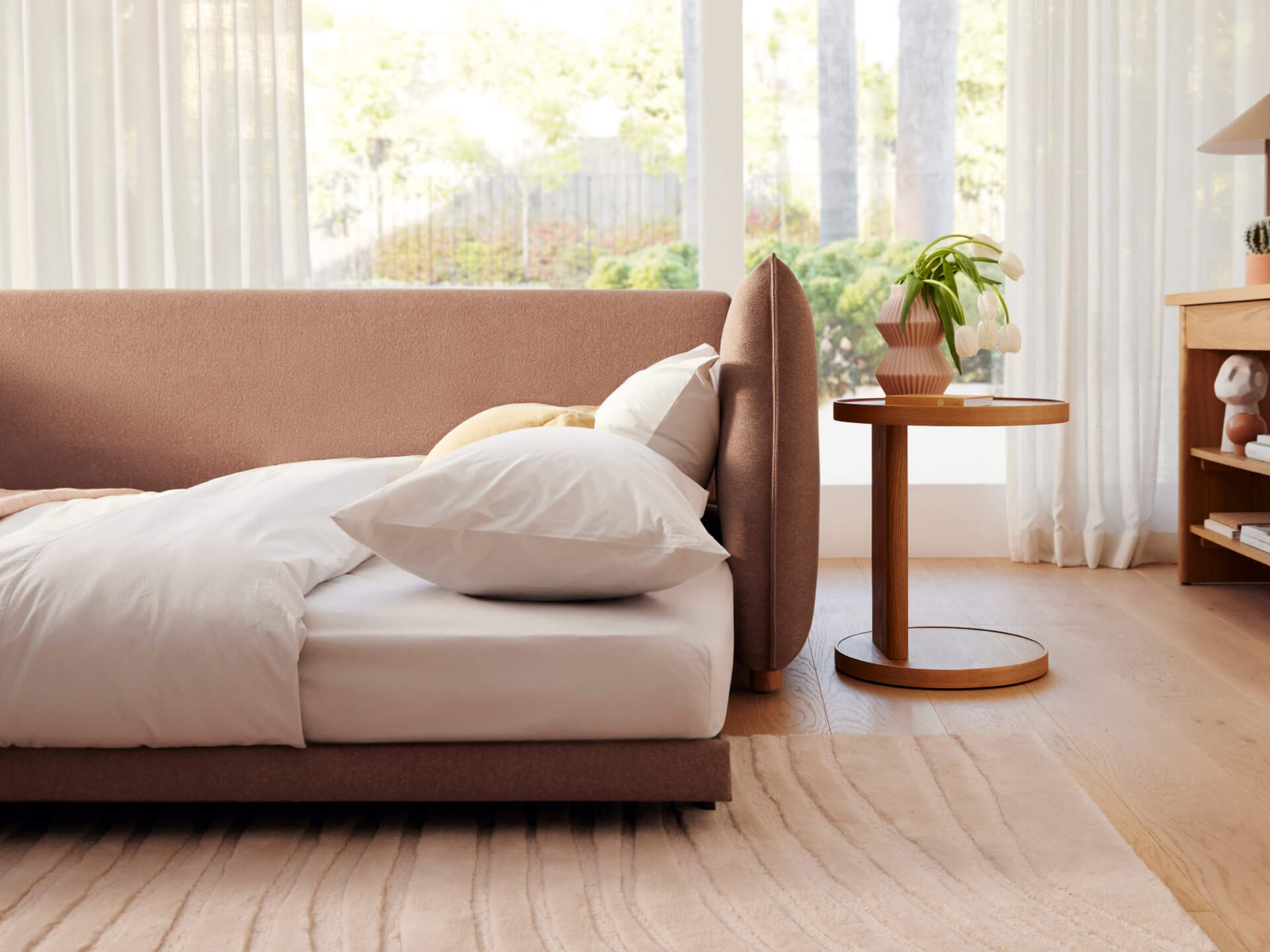 Brown sofa bed with white bedding and pillows, set in a well-lit room. Wooden side table with a plant, light curtains, and a rug complete the decor.