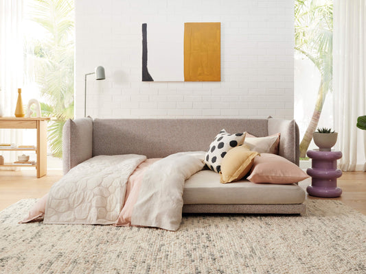 A light grey sofa bed with pastel bedding and assorted pillows on a textured rug. Nearby are a purple side table and a wooden desk against a white wall.