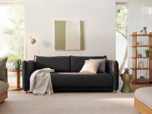 Dark grey sofa with cushions and a throw, against a white brick wall. Wooden shelves and side tables, natural light, minimal decor.