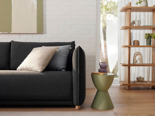 Dark grey sofa with beige pillow, next to a green side table. Wooden shelves with decor in modern living room, white brick wall, and abstract art.