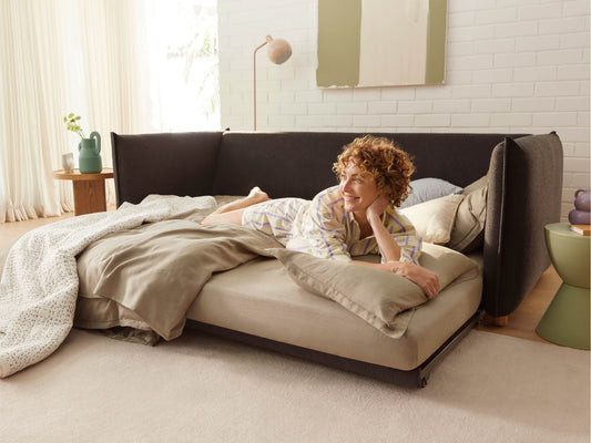 Woman lounging on a light brown bed with grey cushions, surrounded by cosy beige bedding, near a dark grey sofa and green side table. Bright room.