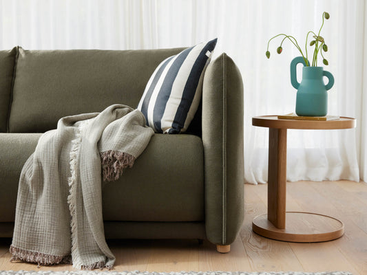 Green sofa with a striped cushion and grey throw, next to a round wooden side table with a teal vase, on light wooden flooring.