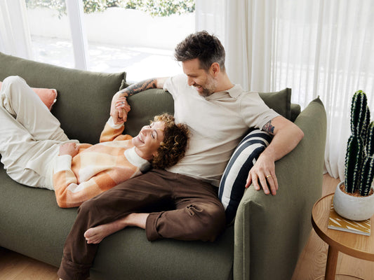 Couple relaxing on a green couch, holding hands. A round wooden side table with a potted cactus is beside them. Natural light through curtains.