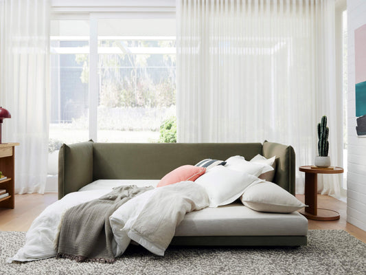 Olive green daybed with white and pink pillows, grey throw, beside a wooden side table with a cactus, in a sunlit room with sheer curtains.