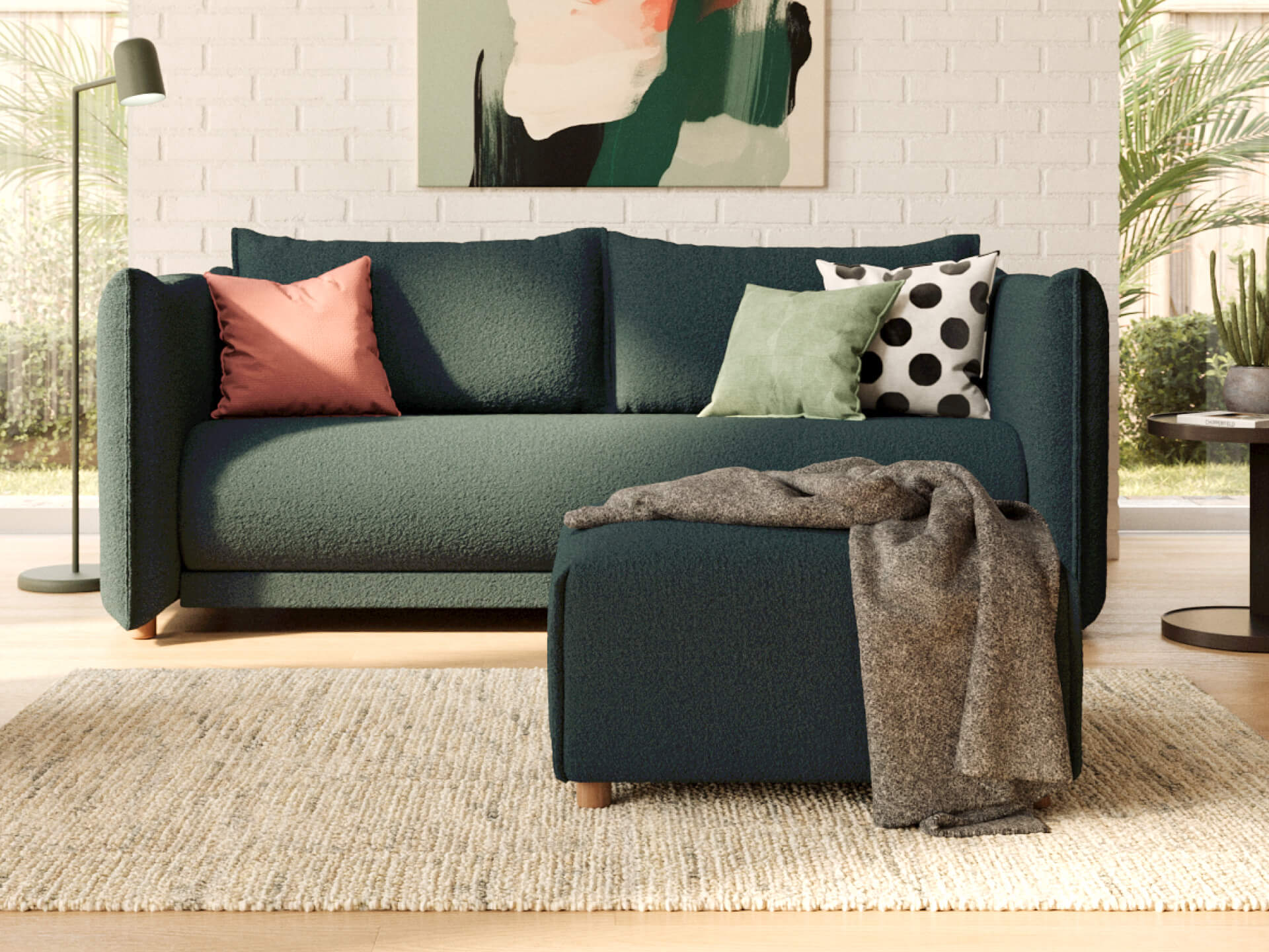 Dark green sofa with colourful cushions; pink, green, polka dot. Matched ottoman, grey throw. Light rug, modern art, and a white brick wall backdrop.