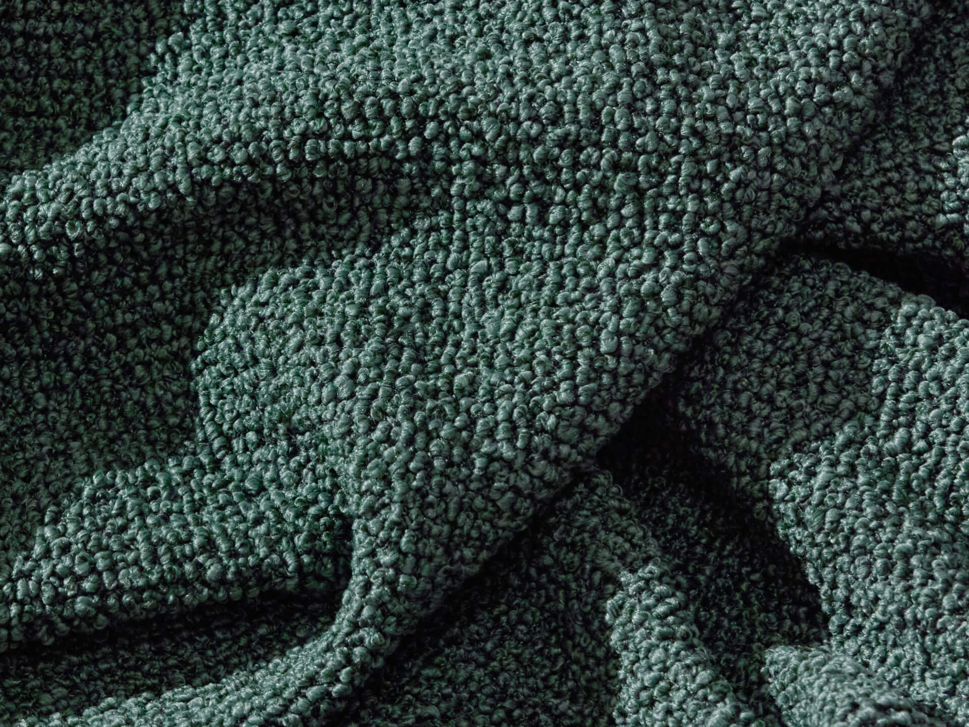 Close-up of dark green boucle fabric with a textured, looped surface pattern, showing folds and shadows. Ideal for upholstery or sofas.