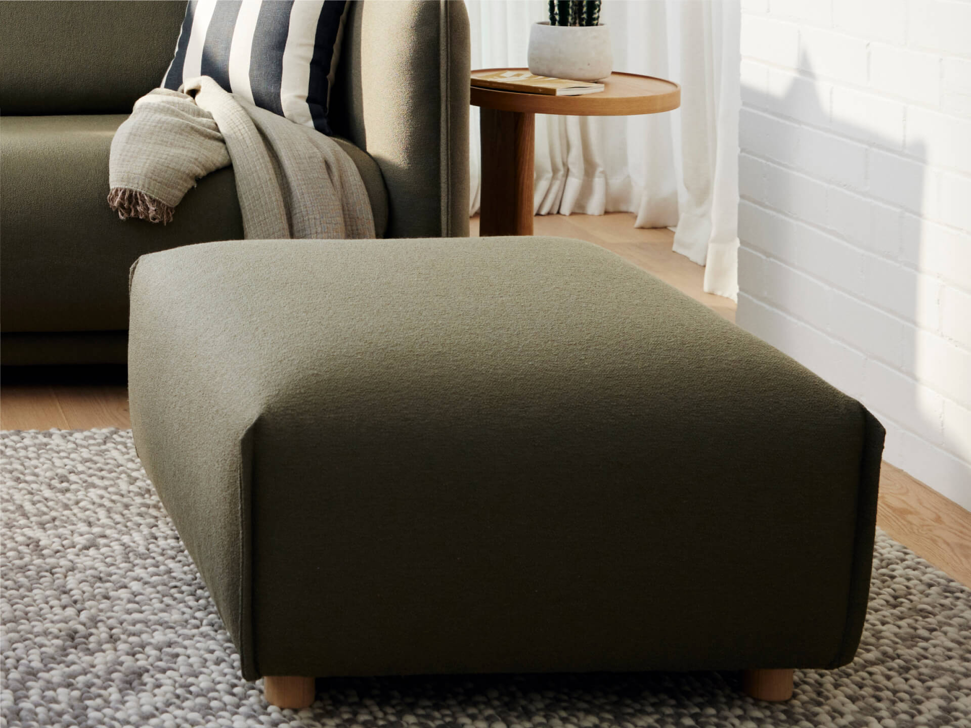 Dark green ottoman on a grey rug, next to a sofa with striped cushions and a draped blanket. Wooden side table with decor is in the background.