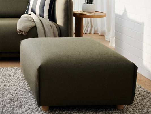 Dark green ottoman on a grey rug, next to a sofa with striped cushions and a draped blanket. Wooden side table with decor is in the background.